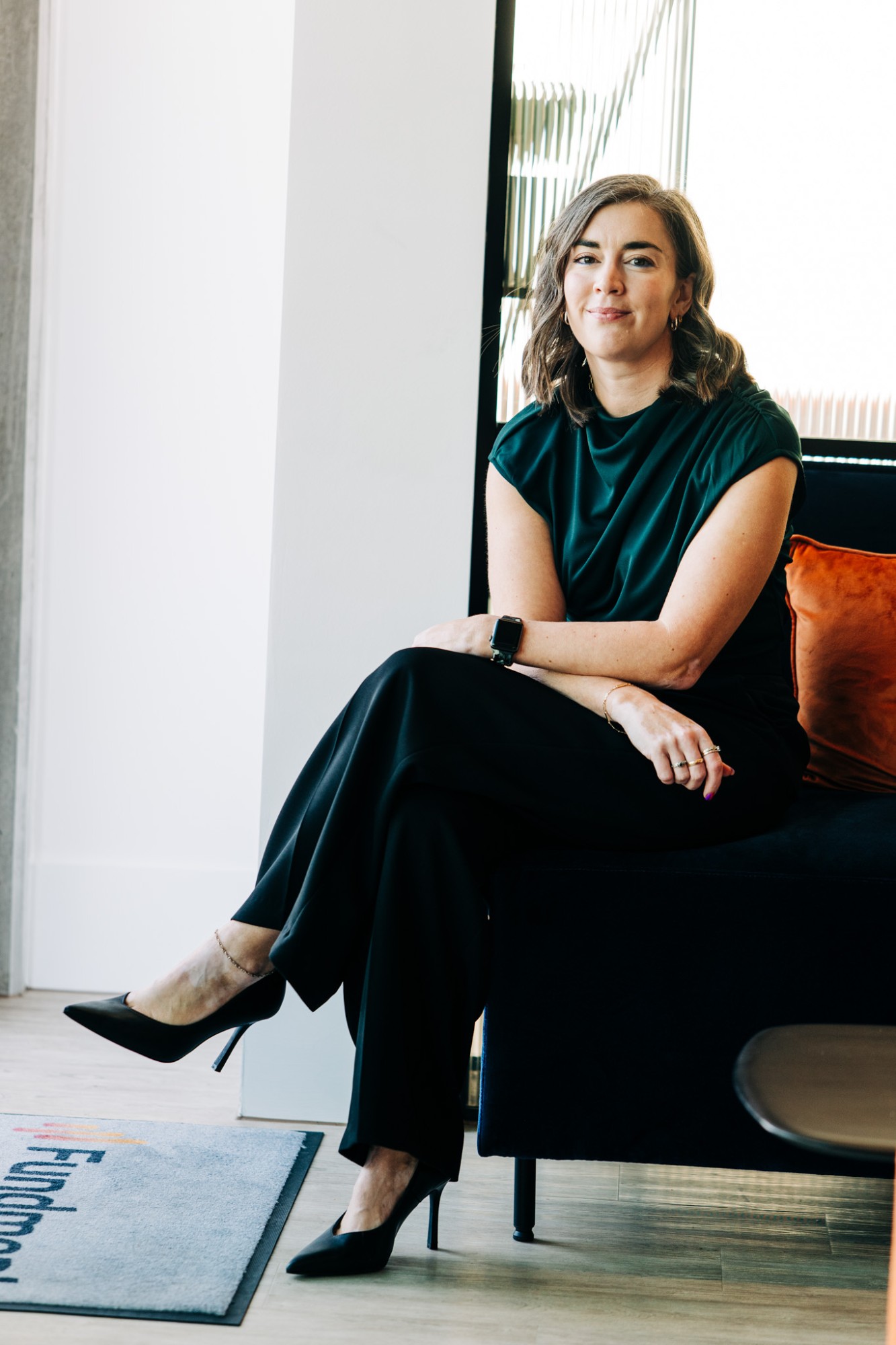 Jancis Dunn seated on a dark sofa by a window, legs crossed, smiling